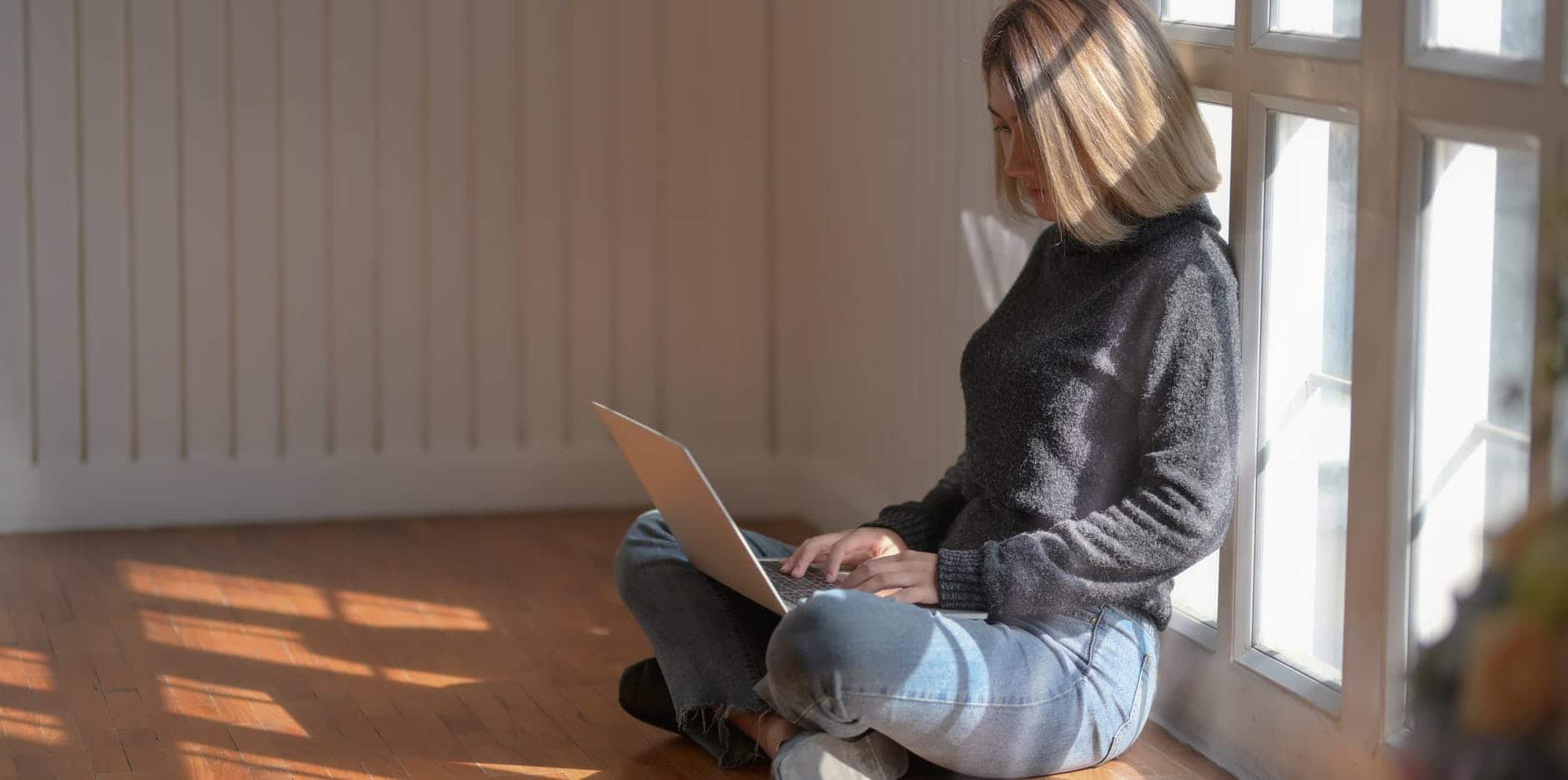 woman in gray sweater using laptop beside glass window